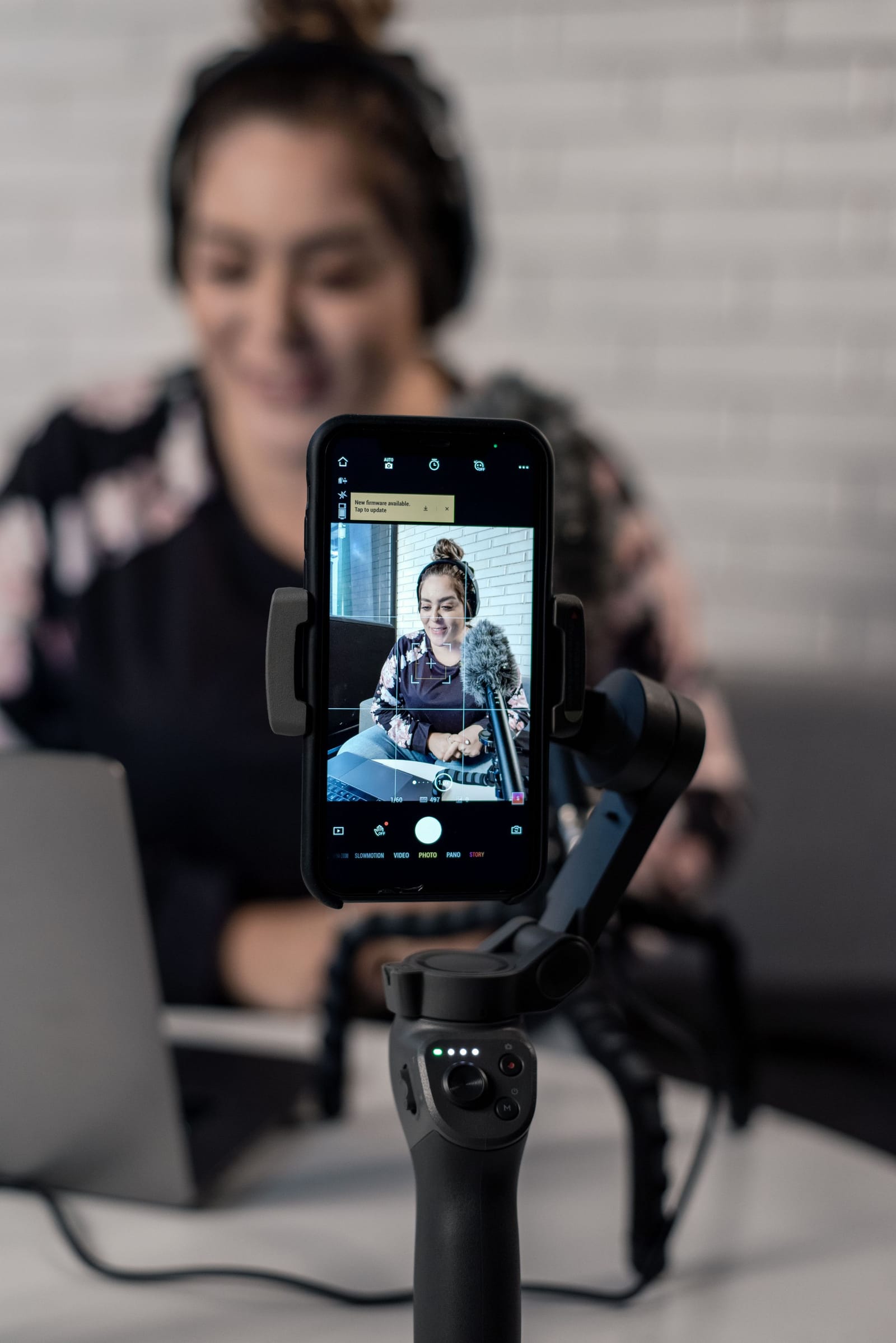Celular no estabilizador grava uma mulher de fone de ouvido falando ao microfone, à mesa com o notebook