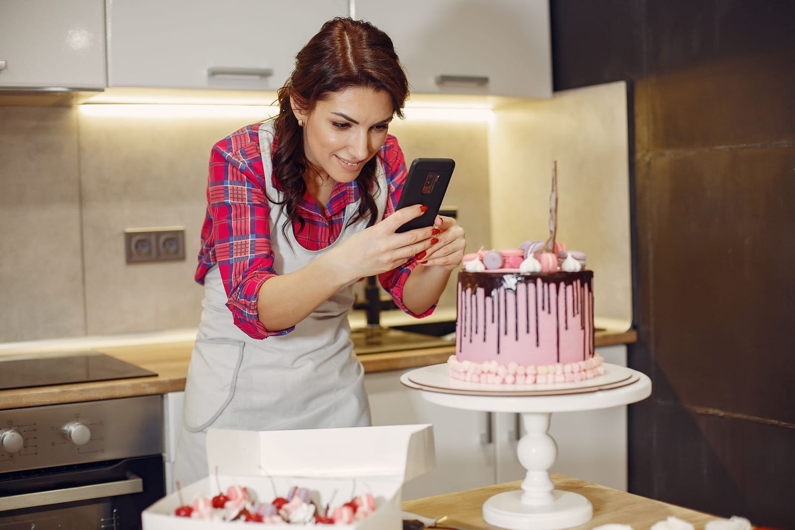 Confeiteira de avental fotografa com o celular um bolo com cobertura de chocolate e macarons