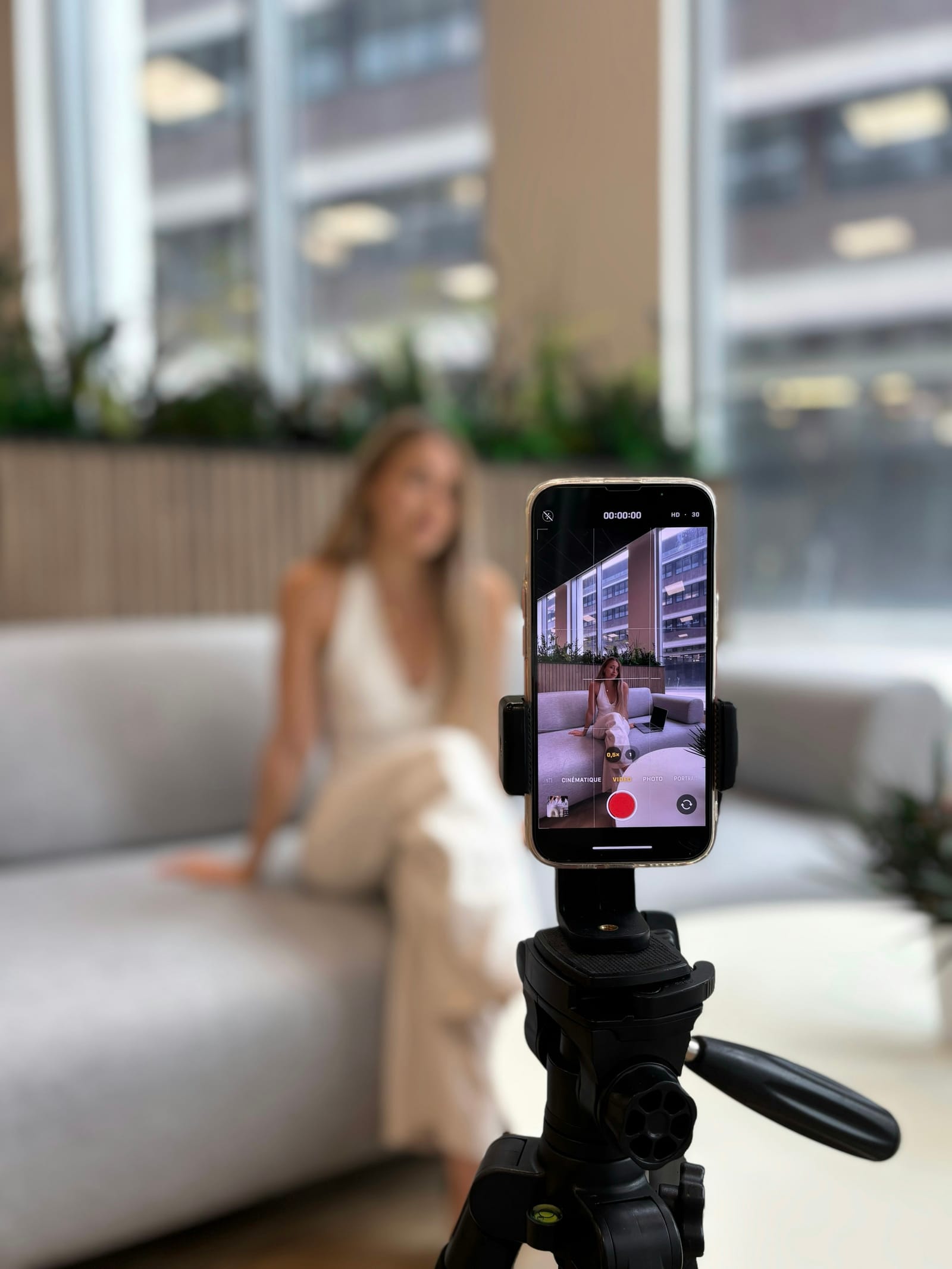 Celular no tripé grava uma mulher sentada no sofá de um escritório; na tela, a gravação em andamento