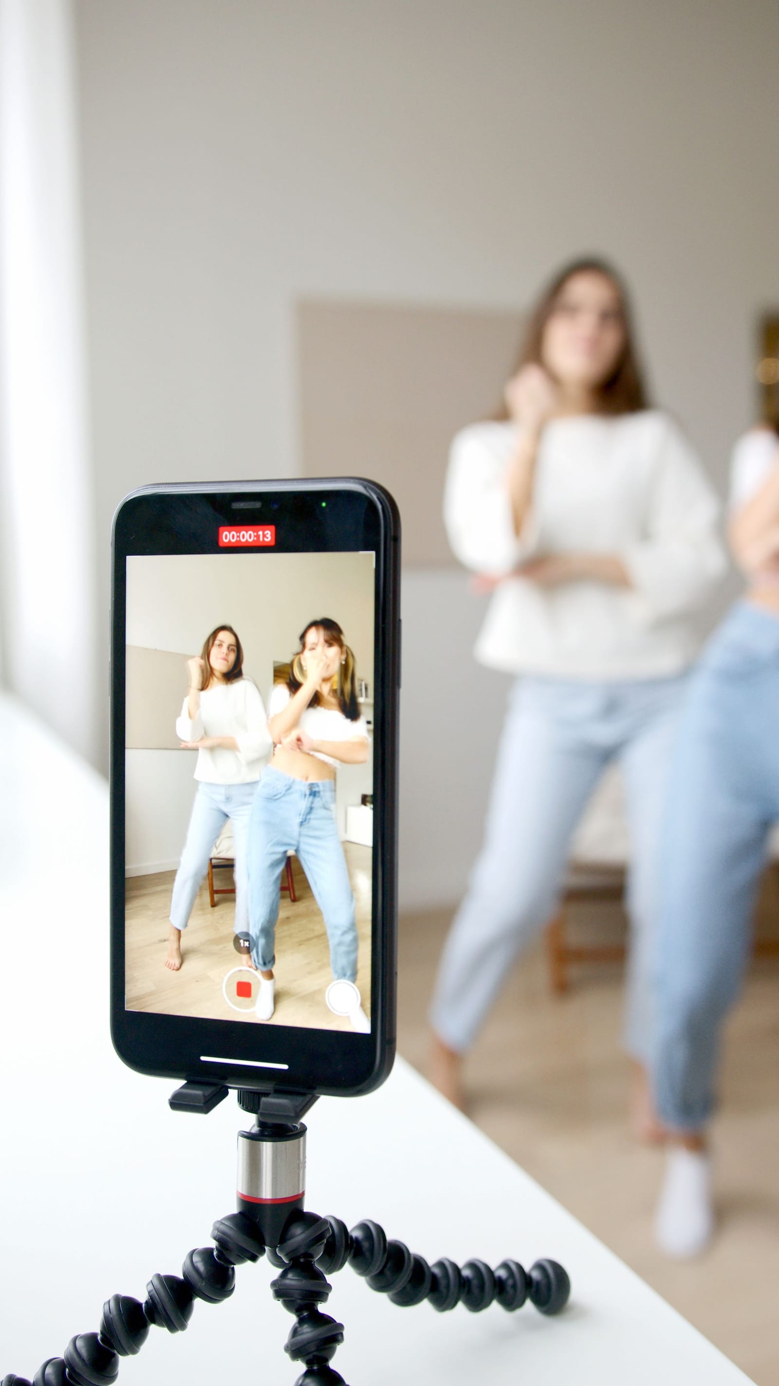 Celular num tripé de mesa grava duas jovens dançando; na tela, as duas aparecem durante a gravação