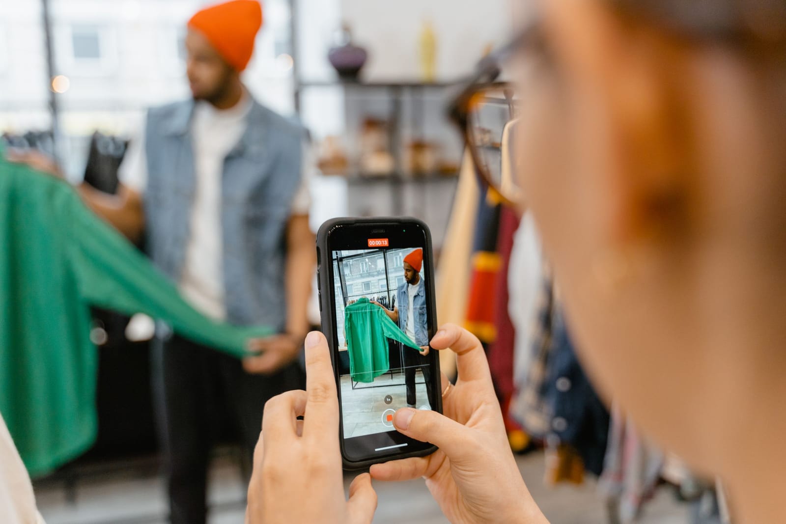 Mãos seguram o celular que grava um vendedor de gorro laranja mostrando uma jaqueta na loja