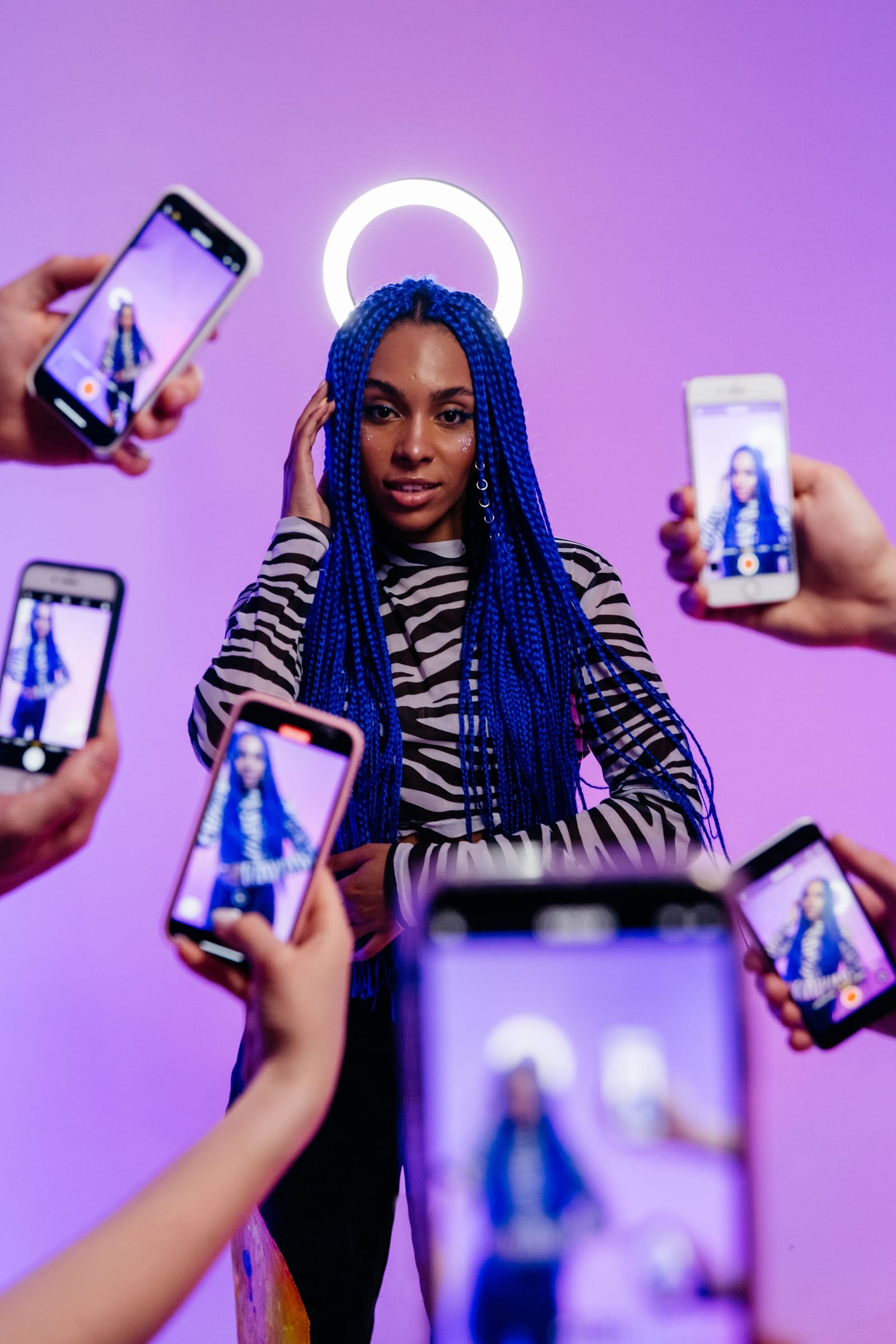 Criadora de tranças azuis posa diante do ring light enquanto várias mãos a filmam com celulares