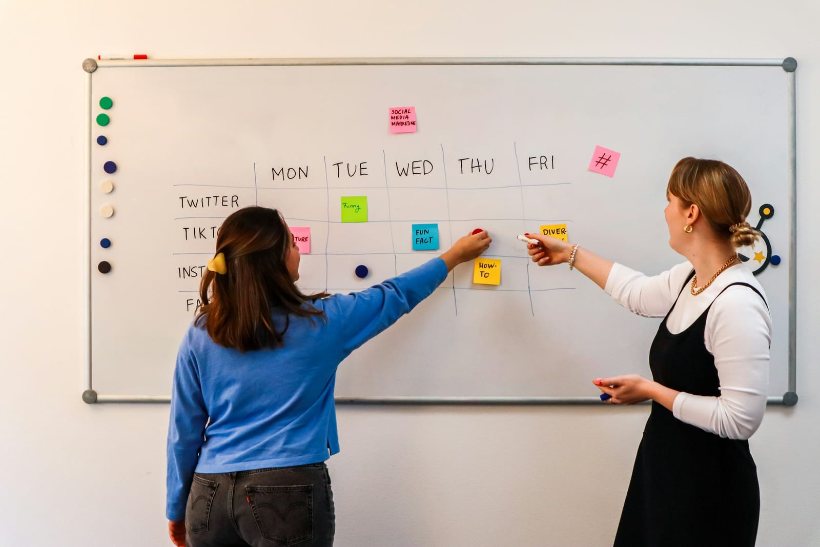 Duas profissionais montam no quadro branco o calendário semanal de conteúdo com post-its
